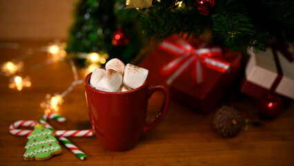 A cup of hot chocolate with marshmallow on wooden table with Christmas tree and decor.