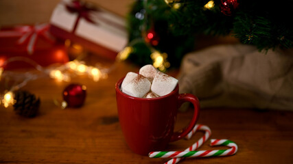 A cup of hot chocolate with melting marshmallow on wooden table with Christmas tree and decor