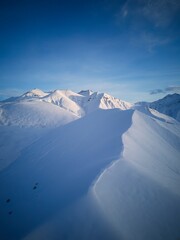 Aerial of snowy mountain slopes full of powder for freeride at ski resort on winter sunrise. Mountains range of backcountry covered with virgin snow. Caucasus peaks skyline with twilight afterglow.
