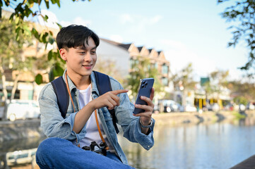 Happy Asian male tourist using his smartphone while relaxes sitting by the small canal.