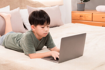Selective focus Asian little child boy laying down on bed using laptop playing games, watching movie, favorite manga or searching internet with curious alone happy at home, natural light