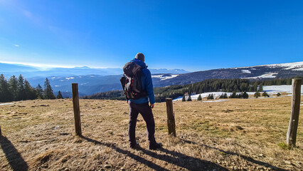 Fototapeta na wymiar Man with backpack on hiking trail standing next to fence on alpine meadow, Saualpe, Lavanttal Alps, Carinthia, Austria, Europe. Trekking on sunny early spring day. View on valley of Wolfsberg. Freedom