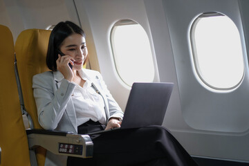Asian woman passenger sitting in airplane near window and reading news from social networks or using travel applications in smartphone