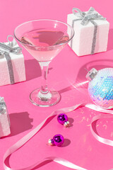 Creative celebration concept with Martini glass, gifts, xmas decorations on bright pink background. Trendy Christmas or New Year party concept.
