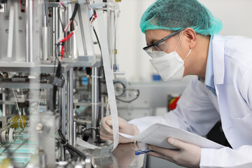 Worker in personal protective equipment or PPE inspecting quality of mask and medical face mask production line in factory, manufacturing industry and fac