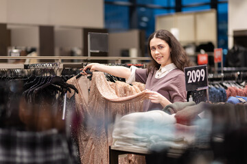 Shopping and sales. Smiling young Caucasian woman showing hanger with sparkly dress. Concept of consumerism and black friday