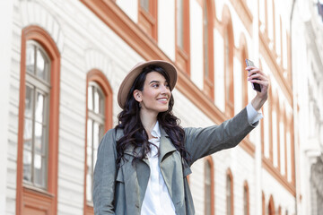 Carefree and happy, sunny spring mood. Cute young smiling girl is making selfie on a camera while walking outdoors. She is wearing casual outfit