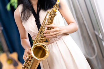 Obraz premium Young woman saxophonist with an instrument stands by the window. Cozy home interior. Concept of hobbies, inner world, development of talents, music school.