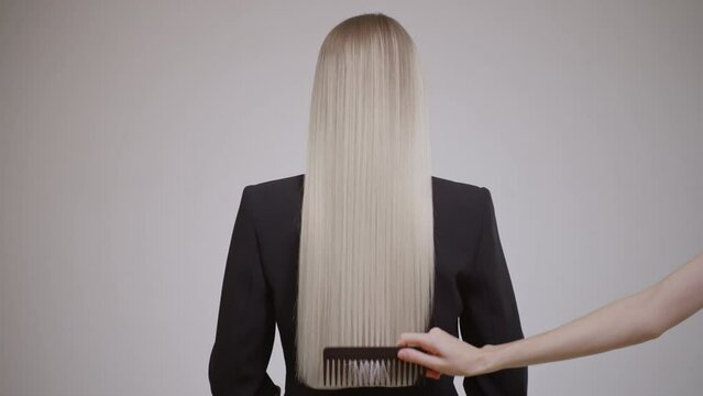 The hairdresser combs the long hair of a blonde woman, rear view. Grey background.