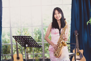 Young woman saxophonist with an instrument stands by the window. Cozy home interior. Concept of...