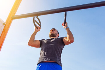 Man in a black t-shirt pulls up on gymnastic rings outdoors, exercising on the sports ground.