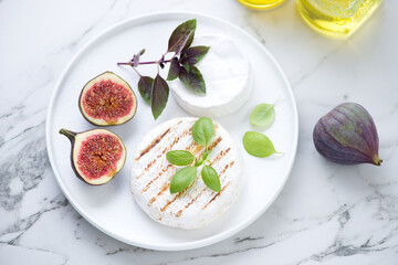 White plate with bbq camembert cheese, fresh figs fruits and basil, high angle view on a light-grey...
