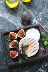 Wooden serving tray with grilled camembert cheese, sliced figs and basil, elevated view on a black marble background, vertical shot