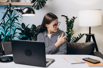 Unhappy young woman in eyeglasses with hand holding shoulder pain. Office syndrome and Health care concept. Businesswoman working with laptop feeling shoulder pain sitting on couch at home office