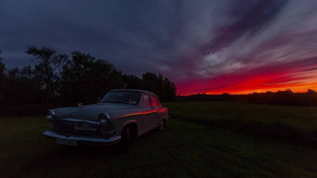 Old historic Gaz 21 car standing on a grassy clearing. In the background, an amazing sunrise in a time lapse shot with a red glow and clouds floating across the sky