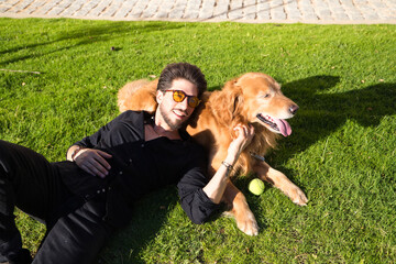 Handsome young man with beard and glasses lying on the grass sunbathing with his brown golden retriever dog. Concept pets, animals, dogs, pet love, travel.