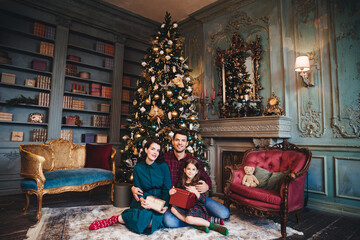 Friendly family sits near decorated Christams tree, embrace each other, hold packed presents, enjoy majestic atmospere. Father, mother and daughter near New Year tree in living room. Festive concept