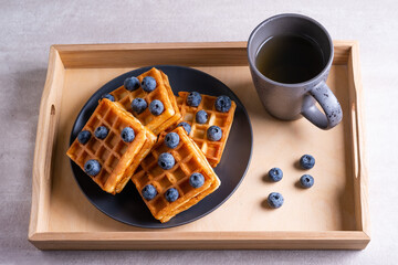 Waffles with blueberries and cup of tea on a wooden tray.