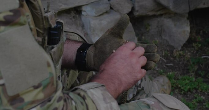 A military man, dressed in a camouflage uniform and tactical gloves, holding a badge and a crucifix on a metal chain in his hand, presses it to his chest, to his heart and praying before the battle.