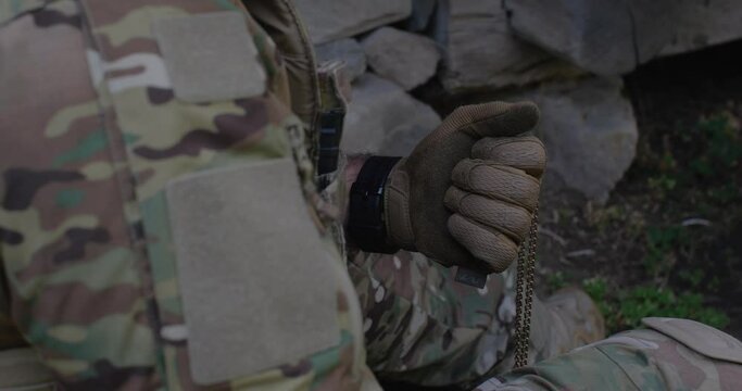 A military man, dressed in a camouflage uniform and tactical gloves, holding a badge and a crucifix on a metal chain in his hand, presses it to his chest, to his heart and praying before the battle.