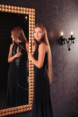 Smiling perfect teen girl in elegant black dress posing at mirror in dark stylish interior room, looking at camera. Cute fashionable teenager lady model showing. Fashion style concept. Copy text space