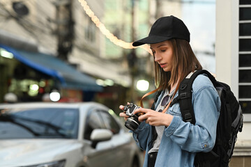 Portrait of pretty young woman traveler wearing hat and backpack standing on busy city street in the evening
