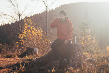 Tourist sitting on a fire pouring hot tea from a thermos into a cup at sunrise. A hiker is...
