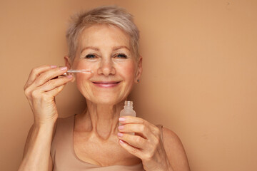 Happy senior lady applies cosmetic oil serum on face takes care of skin and smiles broadly