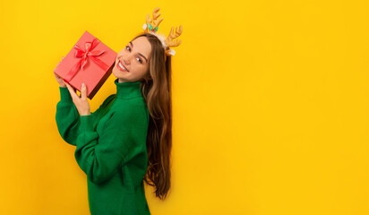 Fototapeta na wymiar Happy smiling cheerful young woman wearing green knitted sweater with Xmas reindeer antlers, holding pink present gift with bow and looking at camera isolated on yellow background with copy space.