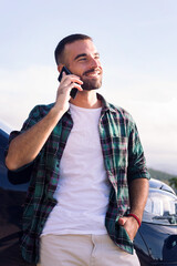 smiling young man talking on mobile phone leaning on his camper van, concept of freedom and nomad...