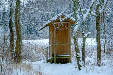 Outdoor wooden toilet house covered with snow on winter forest and trees background. Among the trees by the tourist trail