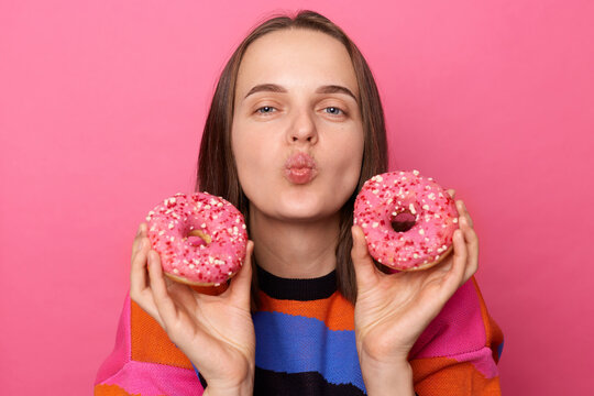 Portrait of attractive romantic flirting woman with dark straight hair wearing jumper standing isolated over pink background, holding glazed tasty donuts and sen ding air kissing.