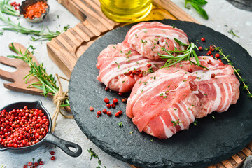Raw cutlets wrapped in bacon served on a black plate with rosemary and thyme. On a stone...