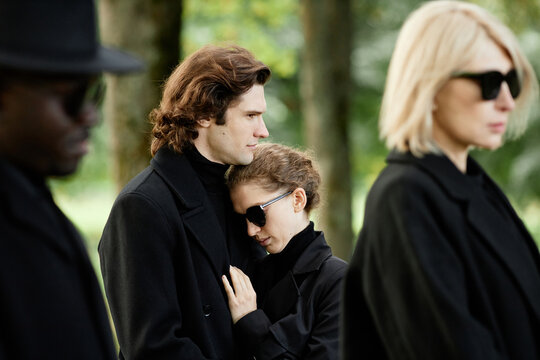 Portrait of young couple wearing black grieving together and embracing at outdoor funeral ceremony - Powered by Adobe