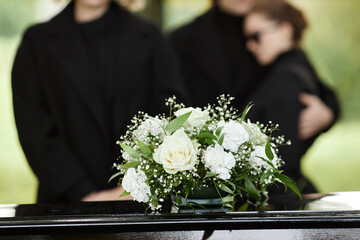 Close up of flower bouquet on coffin with people wearing black in background, outdoor funeral...