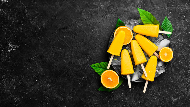 Orange Ice Cream On A Stick. Fresh Oranges And Yellow Popsicles. On A Black Stone Background. Top View.