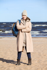 A woman in a coat and with a backpack on her shoulder stands on a sandy beach, arms folded across her chest