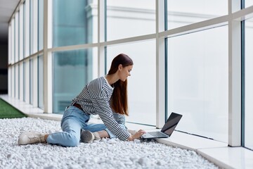 A woman sits in an office lobby by large windows and works on her laptop online via the Internet, freelance work anywhere in the world