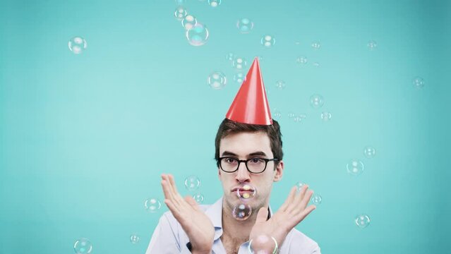 Funny man, bubbles and party hat while dancing, playful and crazy on a blue background to celebrate birthday, holiday or event. Male nerd in photobooth for comic photo at mockup studio party