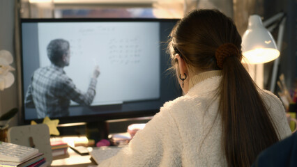 Young asian girl sits at the table at cozy room and learns new material. Schoolgirl listens online lesson of math and studies on pc. Mature teacher talks with student and shows equation on board