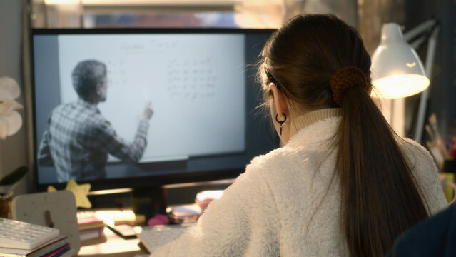 Young asian girl sits at the table at cozy room and learns new material. Schoolgirl listens online lesson of math and studies on pc. Mature teacher talks with student and shows equation on board