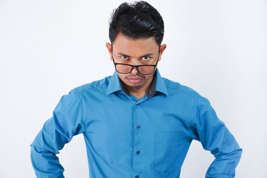 angry asian man with glasses standing with hands on waist and looking at camera wearing blue shirt isolated on white background