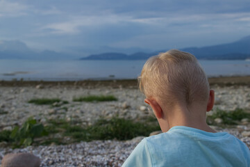 Fototapeta premium A little boy sits with his back on the shore of the lake against the backdrop of blue mountains 