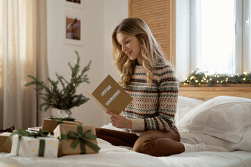 Caucasian woman sitting on bed among Christmas present and reading greeting card