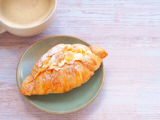 Croissant with cream and almonds on light green plate and coffee cup over wooden table. Close up, space for text..