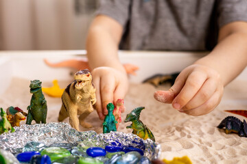 Closeup baby boy hands playing carnivorous and herbivorous dinosaurs with kinetic sand sensory box