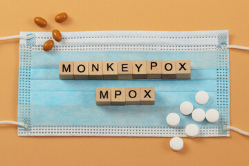 The words Monkeypox and Mpox are laid out with wooden cubes on a surgical face mask. There are various pills lying around. MPOX is the new name for the monkeypox virus.