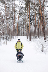 Fototapeta na wymiar Father with backpack and little sos walking together in winter snowy forest. Happy man and joyful boy sledding and having fun together. Wintertime activity outdoors. Concept of local travel