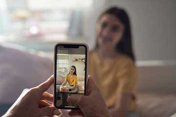Unrecognizable person taking picture with phone of caucasian teenage girl sitting on bed