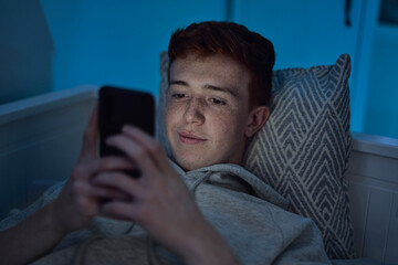 Cheerful caucasian teenage boy using mobile phone while lying in bed at night in his room